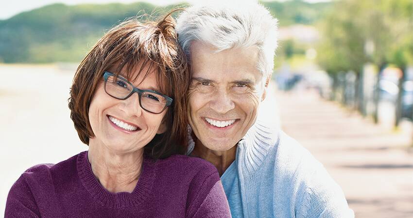Retired couple embraces while smiling to reveal their bright white dental crowns
